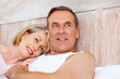 © Stigur/peopleimages.com - Mature couple, portrait and smile on bed at house for relax, love and retirement together with trust and care. Man, woman and happy in hotel room for marriage, loyalty or tired on weekend with hug