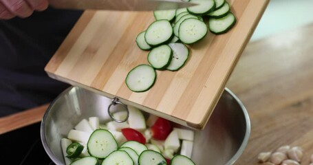 Wall Mural - Man pushes sliced cucumber rings along wooden board with knife pouring into metal bowl. Alternately cutting salad ingredients slow motion