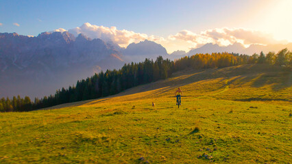 Wall Mural - AERIAL: Fit young lady biking towards picturesque mountaintop with her brown dog