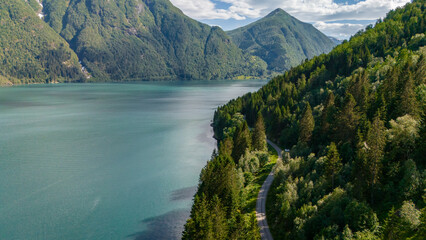 Wall Mural - An aerial view of a winding road through dense forest along the edge of a calm fjord in Norway. The surrounding mountains are covered in lush greenery, Fjaerlandsfjorden, Fjord, Vestland, Norway