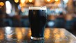 © Dinara - A close-up image of a single glass of dark beer on a bar counter. The glass is filled to the brim with a creamy head of foam.