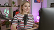© Krakenimages.com - A smiling young woman with blonde hair podcasting with a microphone in a vibrant gaming room lit by a neon cactus sign.