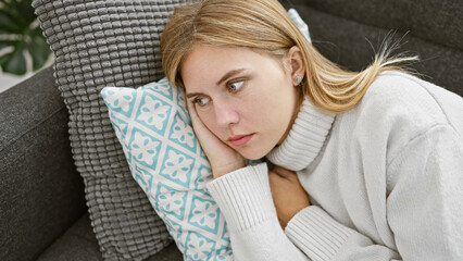 Poster - Pensive blonde woman with blue eyes wearing a sweater, resting in a modern living room on a gray couch with a patterned pillow.