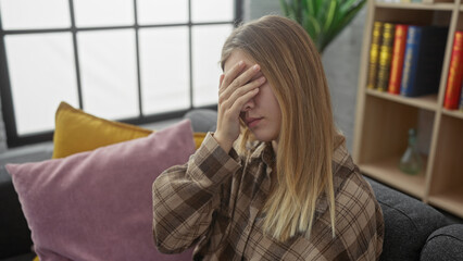 Canvas Print - A young blonde woman indoors covers her eyes with her hand, showing distress against a cozy home background.