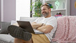 © Krakenimages.com - Hispanic mature man with grey hair smiling while using a laptop on a sofa in a modern living room.