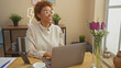 © Krakenimages.com - Smiling african american woman working on laptop at home, with glasses and casual attire in a modern room with plants.