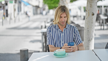 Sticker - A pensive adult man with long blond hair using a smartphone at an outdoor cafe terrace in the city.