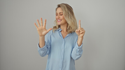 Wall Mural - A smiling young caucasian woman in a blue shirt gestures the number six against a white isolated background.