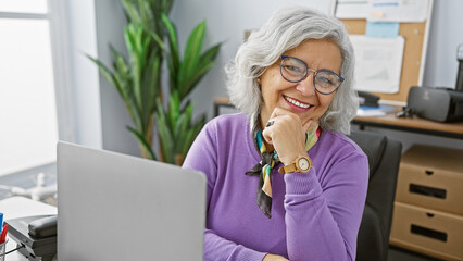 Poster - Smiling mature woman with glasses in a stylish office setting, exuding confidence and professionalism.