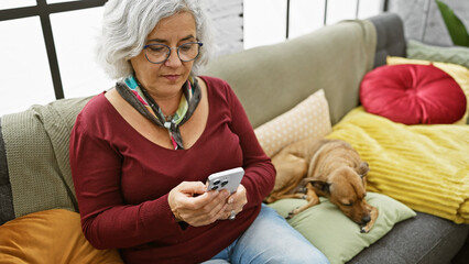 Wall Mural - A mature woman uses smartphone on sofa beside sleeping dog in cozy living room.