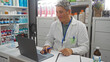 © Krakenimages.com - Pharmacist working on laptop in pharmacy, wearing white coat and badge, surrounded by shelves of medicine and products.