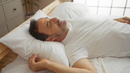 Poster - Hispanic man in white shirt lying on bed in bright bedroom with peaceful expression