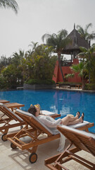 Wall Mural - A young woman relaxes on a sun lounger by a resort pool in bali, absorbing the tranquil tropical ambiance.