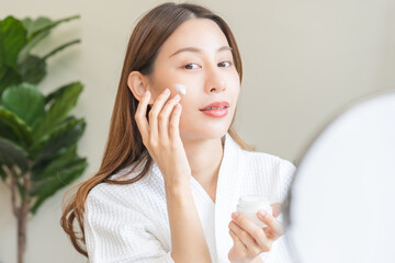 Wall Mural - Facial beauty skin care, smile of pretty asian young woman in bathrobe looking at mirror, hand applying moisturizer lotion on her face, holding jar of skin cream before makeup cosmetic routine at home