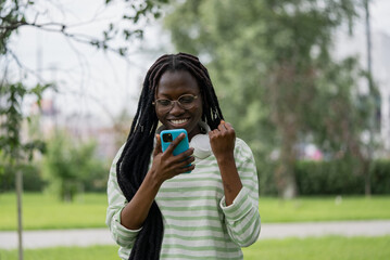 Excited African American woman with headphones celebrates good news while looking at her smartphone in a park, smiling brightly. 