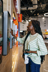 African American girl Using Self-Service Kiosk at Fast Food Restaurant. Black Woman in striped shirt interacting with a self-service kiosk in a store, holding a smartphone and headphones. 