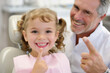 © Elena - Little girl smiling in dentist's chair and pointing finger at her teeth, dentist smiling behind the girl's back, concept of dentistry, caring for health, children's checkup