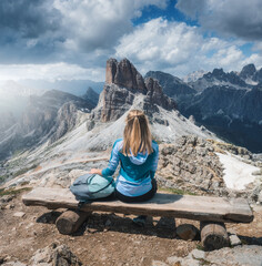 Wall Mural - Girl with backpack is sitting on the wooden bench on mountain peak in beautiful alpine mountains at sunset in summer. Landscape with young woman in alps, high rocks, sky with clouds. Dolomites, Italy