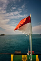 A old tattered Indonesian flag waves in the wind against a serene blue ocean backdrop. Two boats float on the horizon under a partly cloudy sky, showcasing a tranquil maritime scene.