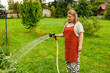 © mdyn - Happy woman wearing apron working outdoors in garden, spraying plants with water from hose. The farmers wife waters the vegetables. The concept of caring for agricultural plants and harvesting.