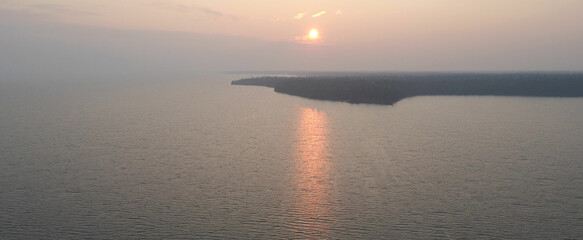 Wall Mural - An aerial view of a very large northern lake that is enveloped in thick forest fire smoke. The distant shoreline is covered in a thick forest. The smoke is creating an ominous orange colored atmospher
