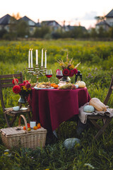 Wall Mural - Beautiful decorated candlelight fall dinner, covered with a red tablecloth with flowers, pumpkins, candles, wine glasses. Outdoor in field near the forest in nature. Wedding banquet, sunset, evening