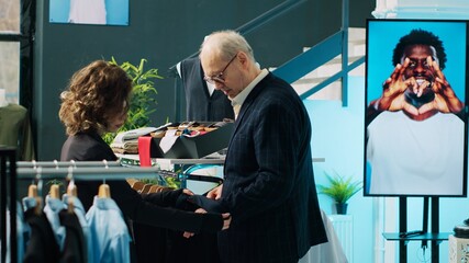 Wall Mural - Woman retail clerk showing multiple tie colors to customer, helping him to match formal suit with the perfect accessories. Employee assisting senior client during shopping spree at mall. Camera A.