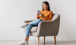 © Prostock-studio - Charming young Asian woman using cellphone, communicating on web, working or learning online, sitting in armchair against white wall, free space. Lovely millennial lady chatting on smartphone