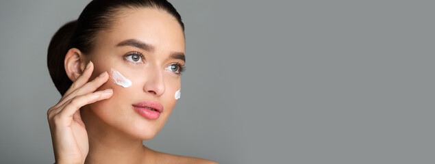 Wall Mural - Beautiful Woman Applying Face Cream and Looking Away, Grey Studio Background, Panorama With Copy Space