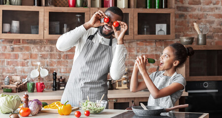 Wall Mural - Afro Man And Little Girl Having Fun At Kitchen, girl making singing in cucumber, dad making tomato eyes, copy space