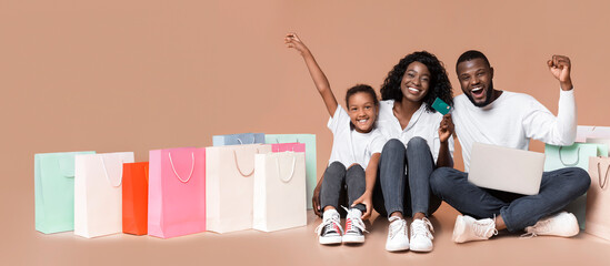 Wall Mural - Online shopping for family. Happy african american parents and daughter sitting on floor with laptop, credit card and bright shopper bags.