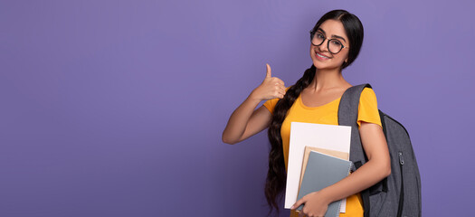 happy indian female student wearing glasses showing thumbs up sign gesture, holding college material