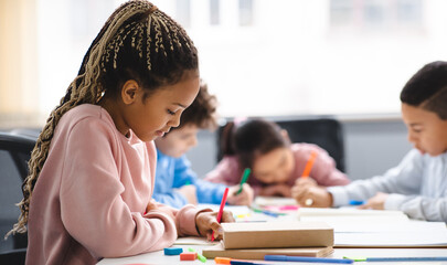 Poster - Portrait of small black girl with afro braids sitting at table in classroom at primary school or kindergarten, writing or drawing in notebook. Reopening and return back to school after lockdown