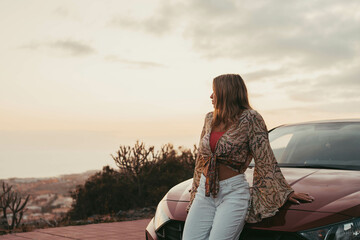 Wall Mural - One young beautiful woman sitting on car outdoor looking at the sunset having fun. Blonde millennial girl feeling good and free at evening..