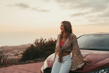 Wall Mural - One young beautiful woman sitting on car outdoor looking at the sunset having fun. Blonde millennial girl feeling good and free at evening..