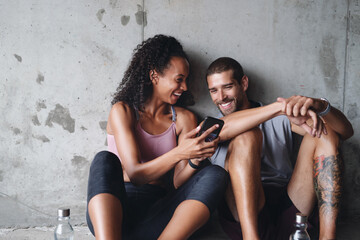 Canvas Print - Man, woman and fitness break with phone for exercise podcast, streaming in building for wellness. Couple, mobile app or workout together with rest in parking lot for health, video for gym or training