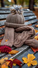 Poster - A brown knitted hat with a pom-pom and a matching scarf are lying on a weathered wooden bench. The bench is surrounded by fallen autumn leaves in shades of yellow, orange, and red.