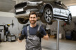 © sofiko14 - Smiling auto mechanic giving thumbs up in car repair shop. Confident technician in uniform standing in front of elevated vehicle in garage. Professional car maintenance and repair service.