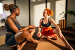 © DusanJelicic - Two diverse women sitting on yoga mats in a studio, engaged in conversation. They are wearing athletic wear and appear to be taking a break from a workout or yoga session.