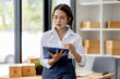 © David - Startup small business entrepreneur SME, asian woman checks stock on clipboard and parcels at home ,Success small business owner online sell marketing delivery, SME e-commerce telemarketing concept