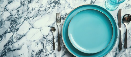 Poster - Trendy dinnerware on a marble background featuring a modern tableware set with cutlery a glass and a vibrant blue plate offering copy space image