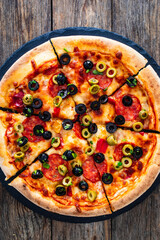 Wall Mural - Circle pizza chorizo with black and green olives and mozzarella served on black stony board on wooden table 