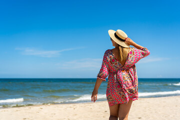 Wall Mural - Beach holidays on Baltic Sea. Young plus size woman wearing fashionable multicolored pink dress with hat standing on sunny sandy beach. Back view