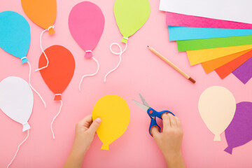 Wall Mural - Little child hands holding scissors and cutting colorful balloon shapes from application paper on pastel pink table background. Making festive decoration. Point of view shot. Closeup. Top down view.