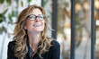 © IBEX.Media - A confident middle-aged Caucasian woman with wavy hair and glasses, smiling brightly in an indoor setting with blurred background