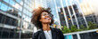 © IBEX.Media - A confident Afro-American businesswoman with curly hair and glasses, smiling and looking upwards while standing in front of modern glass buildings