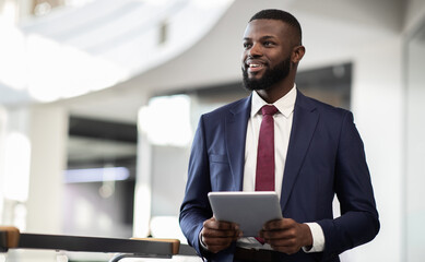 Poster - Cheerful young bearded african american man manager walking by office building corridor, holding modern digital tablet, using mobile app for business, looking at copy space and smiling, panorama