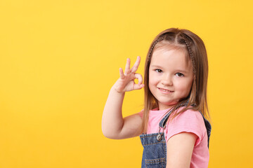 Wall Mural - Portrait of cute little girl showing OK gesture on orange background, space for text