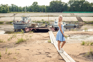 A girl in a blue dress walks along the river bank against the backdrop of old ships.