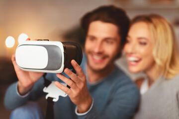 Wall Mural - Cheerful Spouses Holding Virtual Reality Headset Relaxing At Home And Playing Videogame. Selective Focus, Shallow Depth